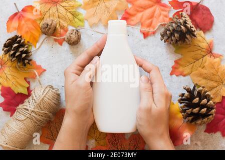 Cosmétiques d'automne, bouteille blanche avec shampooing ou gel douche dans les mains des femmes sur fond gris avec des feuilles jaunes et rouges Banque D'Images