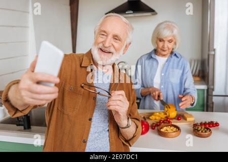 un mari aîné heureux avec un smartphone qui prend le selfie avec une femme qui fait la cuisine dîner dans la cuisine sur un premier plan flou Banque D'Images