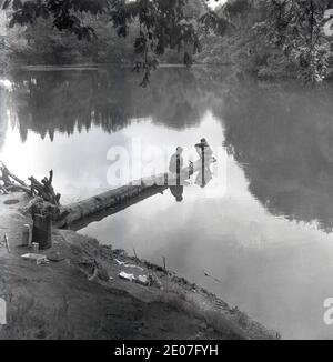 Années 1950, historique, deux jeunes garçons à l'extrémité d'un arbre tombé au milieu d'un lac, utilisant des branches coupées pour pêcher, Angleterre, Royaume-Uni. Banque D'Images