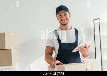 enjoué mogeur en uniforme tenant la tablette numérique près des boîtes dans appartement Banque D'Images