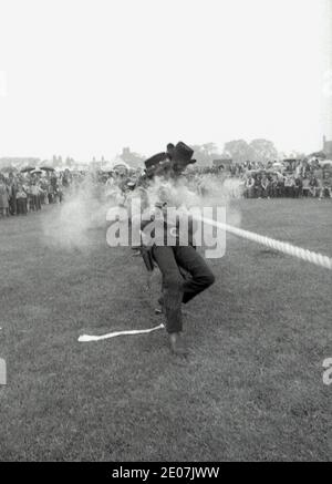 Années 1970, historiques, à l'extérieur d'une tête de village, des hommes en costume de cow-boy se disputant dans un concours de remorqueurs de guerre, et en même temps de tirer des armes dans un événement de démonstration pour divertir la foule, Angleterre, Royaume-Uni. Banque D'Images