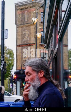 Un homme avec une barbe et des cheveux longs fume la cigarette. En arrière-plan est classé Grade ll bâtiment sur 123 Bethnal Green Rd, à l'angle de Brick Lane, Londres Banque D'Images