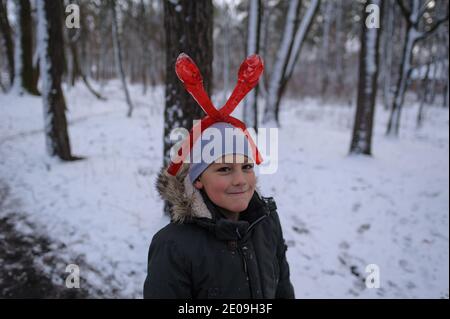 joyeux garçon caucasien dans des vêtements d'hiver vêtu d'une machine à boules de neige sur la tête et souriant dans une forêt d'hiver avec neige Banque D'Images