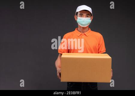 Livraison un homme asiatique ou un coursier portant un uniforme orange masqué tient une boîte en carton dans le studio de concept de coronavirus. Banque D'Images
