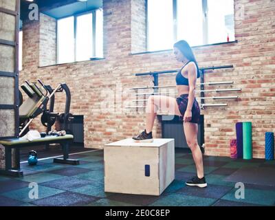 Concept d'entraînement. Brunette femme en vêtements de sport se préparant à sauter sur la boîte en bois dans la salle de gym. Formation fonctionnelle Banque D'Images