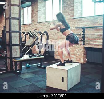 Concept d'entraînement. Une femme brune attirante dans les vêtements de sport saute sur une boîte en bois dans la salle de gym. Vue arrière. Formation fonctionnelle Banque D'Images