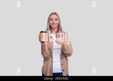 Fille tenant une tasse à café à emporter montrant le pouce vers le haut. Une fille avec une tasse à café à emporter entre les mains. Banque D'Images