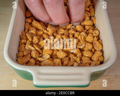 Gros plan POV de tête de cacahuètes torréfiées et enrobées sèches dans un plat en céramique / bol sur une table, avec les doigts d'un homme en prenant certains. Banque D'Images