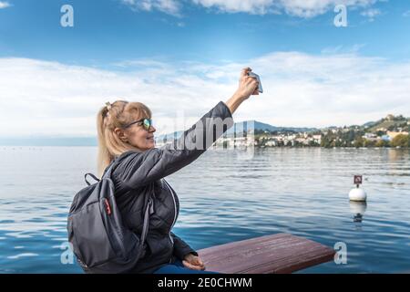 Femme blonde avec des lunettes de soleil et un sac à dos faisant un selfie assis sur les rives d'un lac avec le village et les montagnes en arrière-plan. Mont Banque D'Images