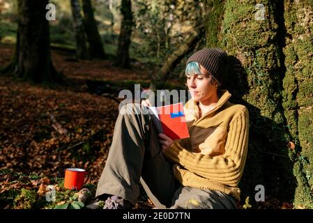 Une femme paisible se penche sur le tronc d'arbre et écrit journal tout en se relaxant dans les bois pendant le voyage d'été Banque D'Images