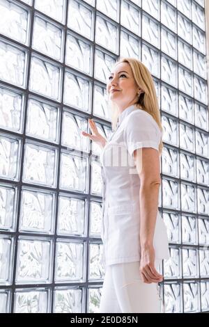 Vue latérale à angle bas de la femme médique en uniforme blanc debout près du mur de verre dans la clinique moderne et regardant loin Banque D'Images