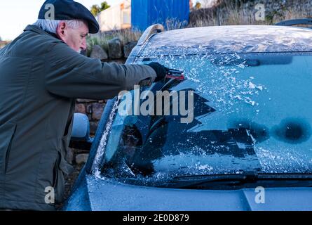 Homme senior raclant la glace du pare-brise de voiture en hiver avec des motifs de gel sur le toit de voiture, Écosse, Royaume-Uni Banque D'Images