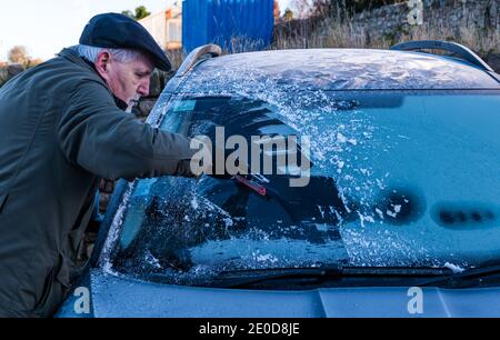 Homme senior raclant la glace du pare-brise de voiture en hiver avec des motifs de gel sur le toit de voiture, Écosse, Royaume-Uni Banque D'Images