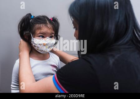 Mère aimante ethnique mettant un masque protecteur sur le visage de mignon petit enfant debout sur fond gris et regardant chacun autre pendant le coronavirus Banque D'Images