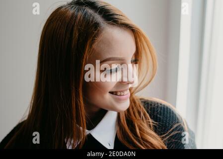 Jeune femme positive avec de longs cheveux rouges et une peau parfaite sincèrement souriant et regardant loin Banque D'Images