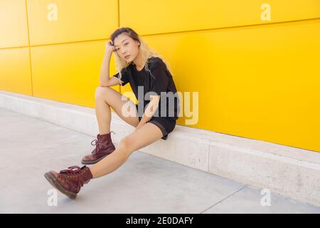 Vue latérale complète de l'asiatique de l'adolescence femme hipster dans vêtements décontractés et bottes près du mur jaune de la ville rue Banque D'Images