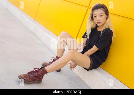 Vue latérale complète de l'asiatique de l'adolescence femme hipster dans vêtements décontractés et bottes près du mur jaune de la ville rue Banque D'Images
