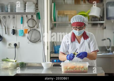 Les femmes cuisent en uniforme blanc et masque et gants de protection couper des légumes frais tout en préparant des aliments dans la cuisine de l'hôpital pendant pande coronavirus Banque D'Images