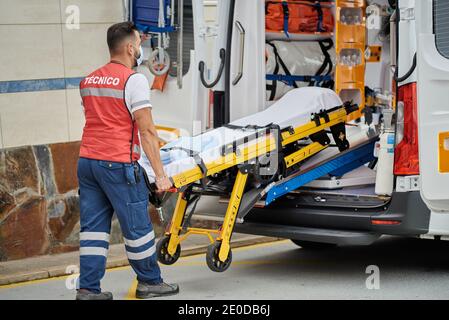 Vue latérale d'un professionnel de l'aide médicale mâle dans le masque de protection tirant civière hors de la camionnette d'ambulance garée dans la rue Banque D'Images