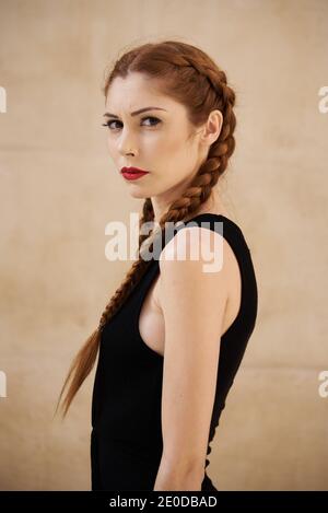 Vue latérale du modèle féminin élégant avec cheveux rouges et tresses assises en studio sur fond sombre et regardant appareil photo Banque D'Images