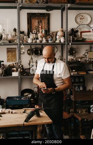 Menuisier d'âge moyen sérieux dans les messages de tablier sur mobile téléphone à proximité d'un établi avec des outils professionnels créatifs studio de travail Banque D'Images