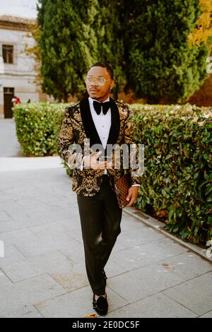 Élégant Afro-américain mâle portant un tuxedo cher marchant le long de la rue et regarder loin Banque D'Images