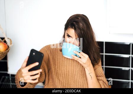 Une femme détendue assise avec une tasse de café sur le comptoir cuisine prenant selfie avec téléphone mobile tout en appréciant le week-end à accueil Banque D'Images
