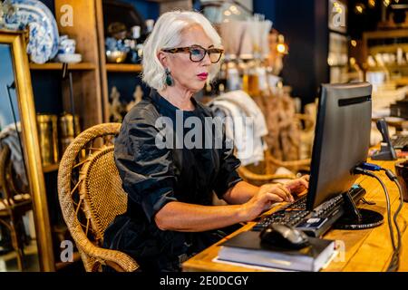 Vue latérale d'une femme d'affaires mature dans une élégante robe assise à la table et en tapant sur le clavier tout en travaillant sur l'ordinateur sur le projet en ligne Banque D'Images