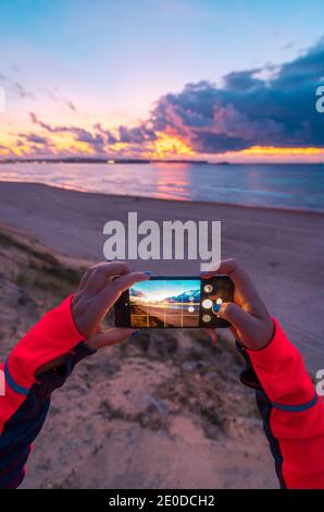 Vue arrière d'une femme voyageur méconnue avec de longs cheveux foncés dans des vêtements décontractés prenant la photo d'un coucher de soleil incroyable sur la mer avec plage de sable sur smartp Banque D'Images