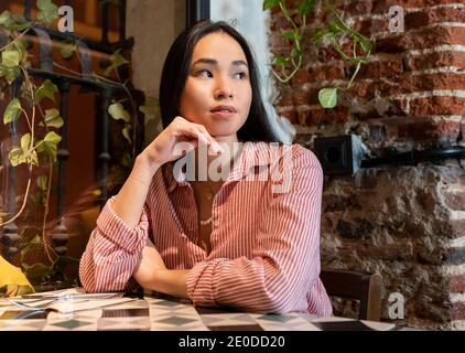 Jeune femme asiatique tranquille qui regarde loin dans des vêtements décontractés assis seul à la table dans le café et en attente de commande Banque D'Images