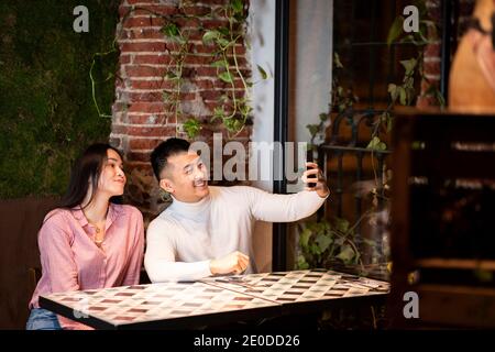 Vue latérale de jeune asiatique gai homme et femme dans des vêtements décontractés assis à la table et prenant le selfie sur smartphone tout en ayant la réunion dans un café confortable Banque D'Images