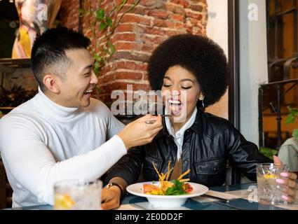 Bon homme asiatique nourrissant une amie afro-américaine joyeuse avec salade tout en s'amusant pendant le déjeuner au café Banque D'Images