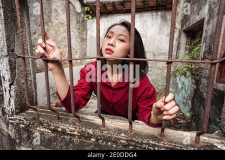 Jeune femme asiatique se tenant à l'intérieur d'un vieux bâtiment en pierre abandonné et regarder la caméra à travers la fenêtre avec la grille métallique rouillée Banque D'Images