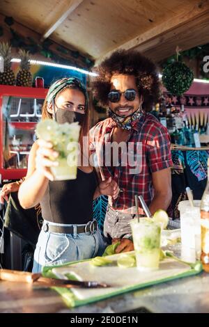 Les propriétaires ethniques positifs du bar tropical regardant l'appareil-photo amical et propose un verre de cocktail rafraîchissant avec du citron vert et de la glace Banque D'Images