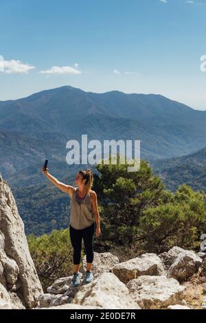 Tout le corps d'une femme exploratrice active se tenant sur une montagne rocheuse en haut et en prenant le selfie sur smartphone contre le paysage montagneux pittoresque en été Banque D'Images