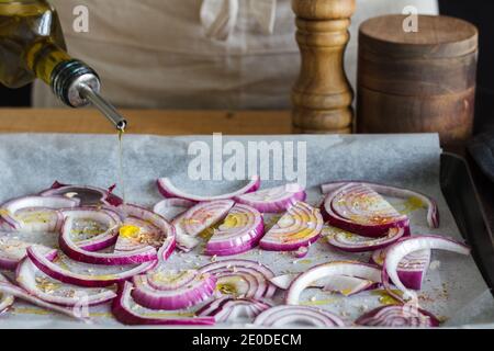 Chef anonyme versant de l'huile d'olive sur des morceaux d'oignons avec assaisonnements placés sur du papier de cuisson sur une casserole dans la cuisine Banque D'Images
