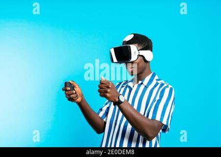 Vue latérale du jeune homme afro-américain dans des lunettes VR et avec joystick, la réalité virtuelle est présente sur fond bleu dans studio moderne Banque D'Images