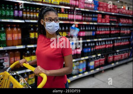 Afro latina jeune femme portant un masque de visage regarde les produits dans l'allée des boissons tout en faisant du shopping dans le supermarché Banque D'Images