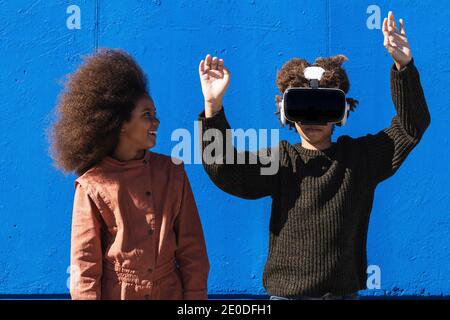 Un garçon afro-américain méconnaissable porte un casque VR et explore le cyberespace tout en se tenant avec une fille contre le mur bleu dans la rue Banque D'Images