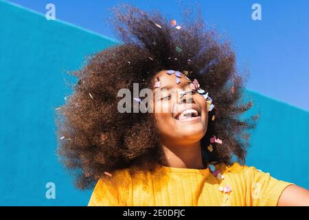 Faible angle de l'adolescence insouciante heureux afro-américaine fille avec longs cheveux bouclés et confettis colorés sur le visage s'amusant contre le mur bleu en été ensoleillé Banque D'Images