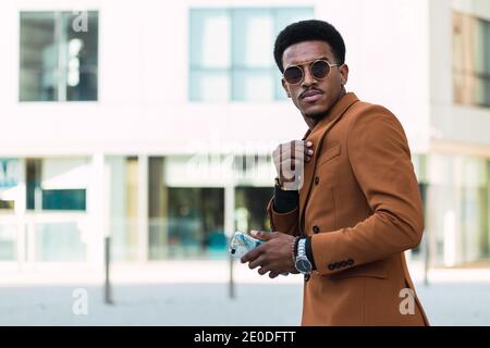 Homme afro-américain auto-assuré portant une veste élégante et des lunettes de soleil réglage du col de la chemise en ville et en regardant à l'appareil photo Banque D'Images