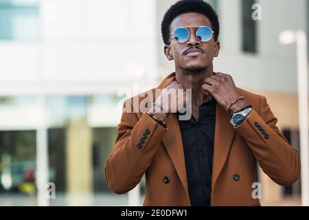 Homme afro-américain auto-assuré portant une veste élégante et des lunettes de soleil réglage du col de la chemise en ville et en regardant à l'appareil photo Banque D'Images