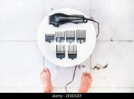 Vue de dessus de Crop anonyme pieds nus homme debout devant de table avec tondeuse à cheveux électrique et accessoires de divers pour couper les cheveux Banque D'Images