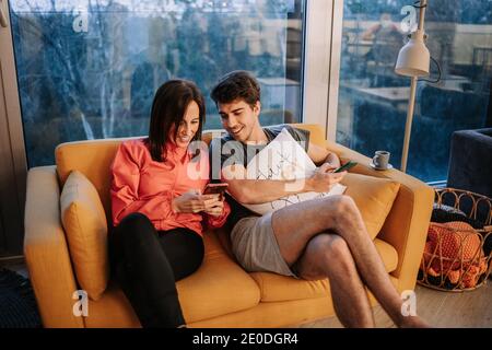 Des amis gais, hommes et femmes, se reposant sur un canapé à la maison et regarder des vidéos sur un téléphone portable ensemble Banque D'Images
