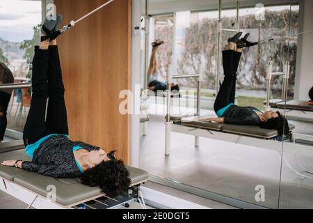 Vue latérale de la compagnie de personnes dans les vêtements d'activité couché Reformeur pilates avec sangles en position de pont avec jambe relevée et faire du yoga en salle de sport ensemble Banque D'Images