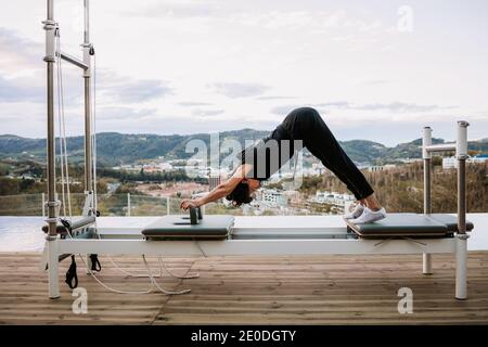 Vue latérale d'une femme gracieuse faisant du yoga à Eka Pada Adho Mukha Svanasana sur les pilates reformer sur la terrasse au coucher du soleil Banque D'Images