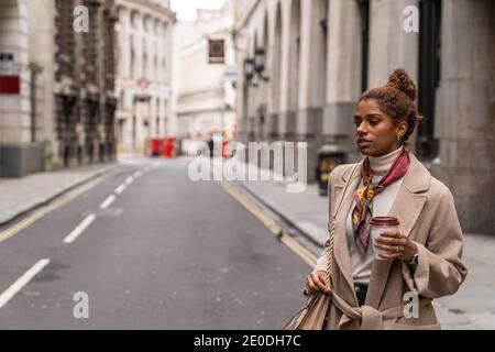 Jeune femme afro-américaine gaie avec des cheveux habillés avec style chemise colorée se tenant près d'un mur de verre et naviguant sur mobile téléphone à l'écart Banque D'Images