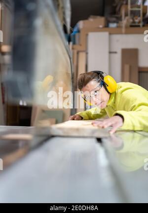 Vue latérale de l'artisan adulte ciblé dans les lunettes et casque scie le panneau en bois avec une scie circulaire dans l'atelier Banque D'Images