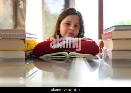 Jolie jeune fille diligente assise à la table, penchée sur les bras à proximité pile de manuels et de regarder l'appareil photo tout en faisant des devoirs cession Banque D'Images