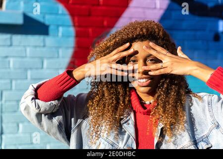 Ravie jeune femme afro-américaine avec de longs cheveux bouclés dedans ensemble décontracté touchant le visage et regardant l'appareil photo contre coloré mur avec graffiti Banque D'Images